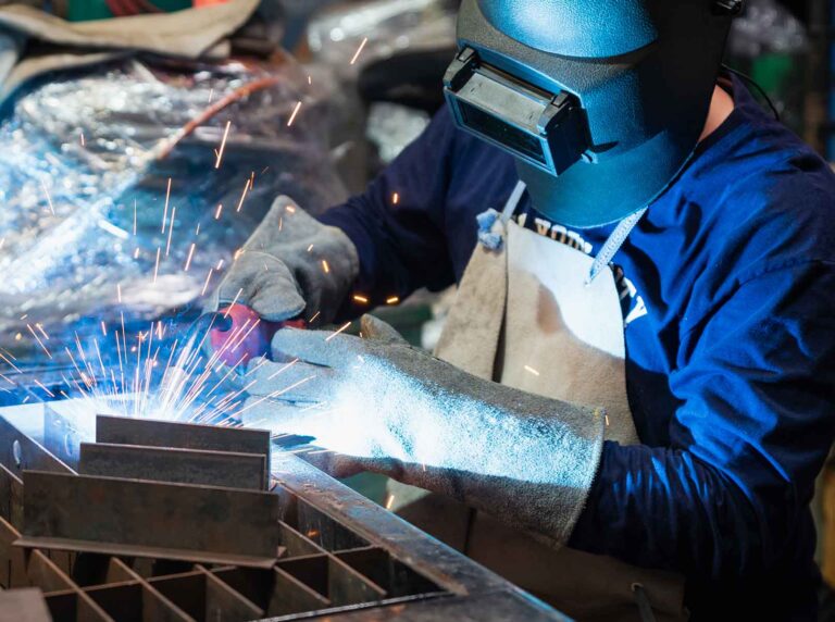 Soldador profissional usando, máscara de solda, luva de raspa e avental de raspa para se proteger das faíscas da solda em uma bancada de trabalho.