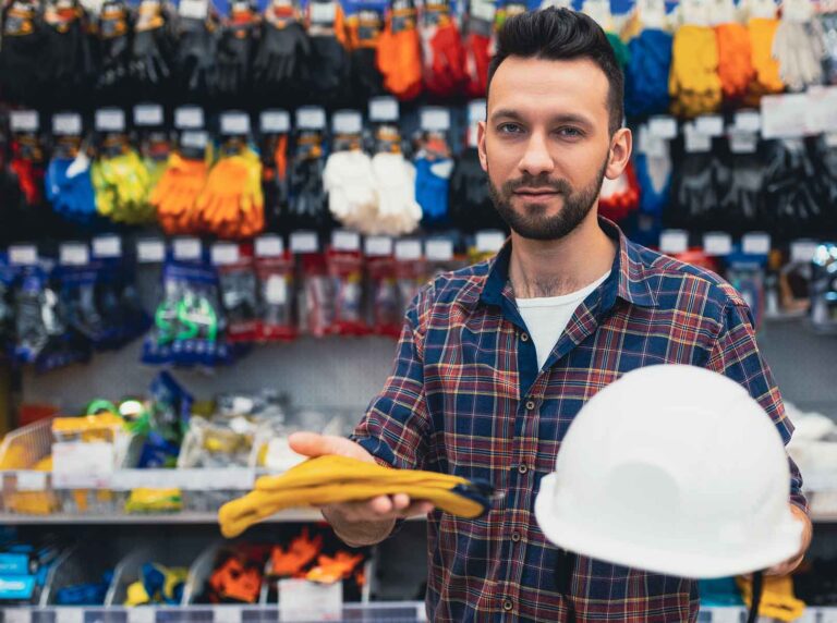 homem-em-frente-as-prateleiras-de-uma-loja-de-epi-segurando-um-capacete-branco-e-luvas-de-protecao-amarelas