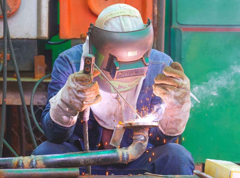 Um operário de solda está trabalhando em um ambiente industrial, usando equipamentos de proteção, como máscara facial, luvas grossas e roupa de trabalho. Ele segura um tubo metálico com uma chama elétrica, gerando faíscas durante o processo de soldagem.