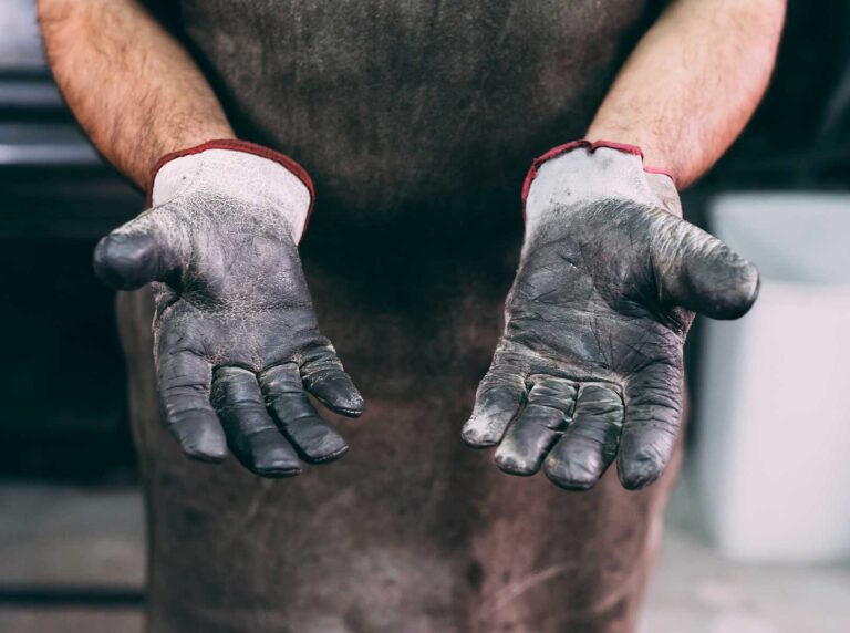 Close-up das mãos de um trabalhador com luvas de vaqueta desgastadas, evidenciando a necessidade diária da proteção das mãos no ambiente de trabalho industrial.