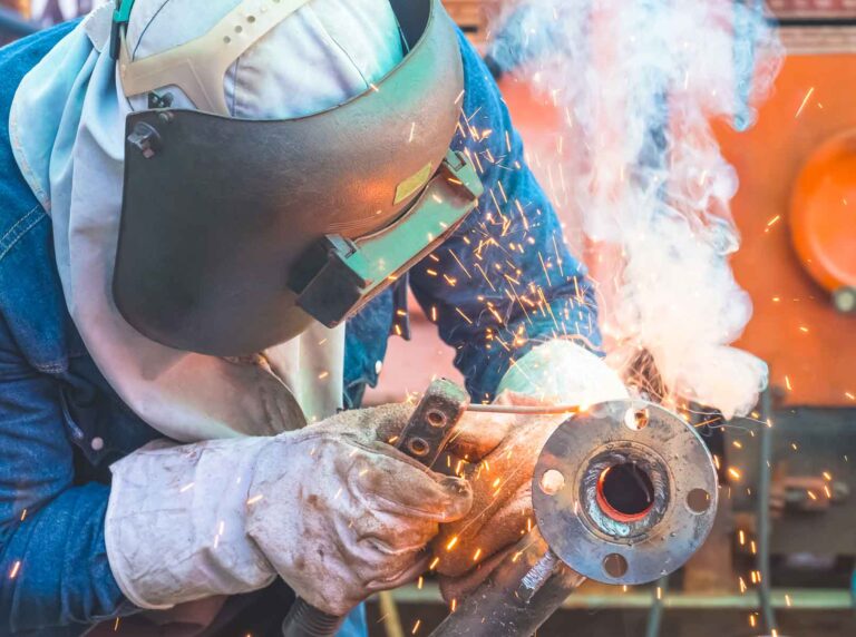 Close de um soldador profissional protegido com máscara de solda gerando uma nuvem de fumos de solda ao unir uma flange metálica.