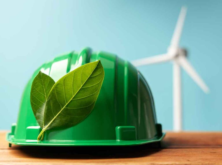 capacete de segurança verde com folhas sobre mesa de madeira e turbina eólica ao fundo representando a integração entre esg e sst