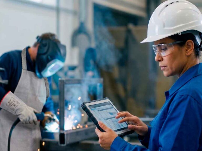 engenheira de segurança do trabalho com capacete e óculos de proteção preenchendo dados em um tablet industrial para o ltcat com um soldador usando avental e luvas de raspa ao fundo