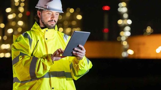 técnico de segurança do trabalho com capacete e jaqueta refletiva usando um tablet para gerenciar as atribuições do técnico de segurança do trabalho em uma planta industrial à noite