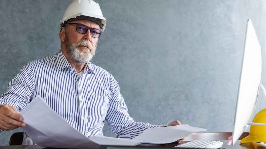 técnico de segurança do trabalho experiente com capacete branco e óculos revisando documentos técnicos em seu escritório. A imagem representa a fase de transição e o desejo de saber como se tornar engenheiro de segurança do trabalho para alcançar cargos estratégicos.