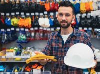 homem-em-frente-as-prateleiras-de-uma-loja-de-epi-segurando-um-capacete-branco-e-luvas-de-protecao-amarelas