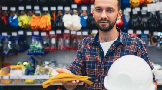 homem-em-frente-as-prateleiras-de-uma-loja-de-epi-segurando-um-capacete-branco-e-luvas-de-protecao-amarelas