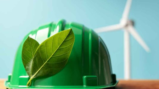capacete de segurança verde com folhas sobre mesa de madeira e turbina eólica ao fundo representando a integração entre esg e sst