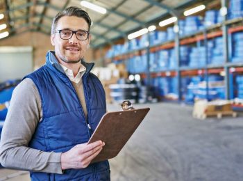 Dono de revenda de EPI no armazém com prancheta, ilustrando a gestão de estoque como parte do guia de negociação.