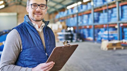 Dono de revenda de EPI no armazém com prancheta, ilustrando a gestão de estoque como parte do guia de negociação.