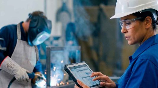 engenheira de segurança do trabalho com capacete e óculos de proteção preenchendo dados em um tablet industrial para o ltcat com um soldador usando avental e luvas de raspa ao fundo