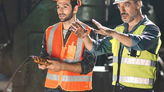 dois profissionais no chão de fábrica. o profissional de sst com capacete branco e colete amarelo orienta o operador que segura um controle, ilustrando a liderança em segurança do trabalho na prática.