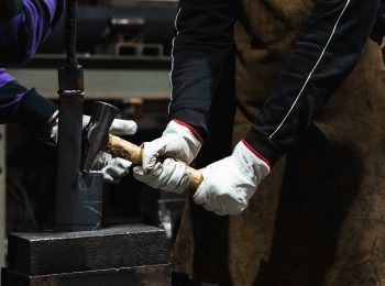 Dois trabalhadores com luvas de vaqueta manuseiam uma ferramenta pesada em ambiente industrial, demonstrando a resistência e o conforto do EPI em tarefas de impacto.