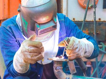 Soldador profissional devidamente equipado com máscara e luva para soldador de raspa, realizando soldagem em uma tubulação industrial.