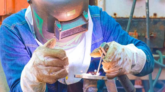 Soldador profissional devidamente equipado com máscara e luva para soldador de raspa, realizando soldagem em uma tubulação industrial.