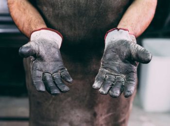 Close-up das mãos de um trabalhador com luvas de vaqueta desgastadas, evidenciando a necessidade diária da proteção das mãos no ambiente de trabalho industrial.