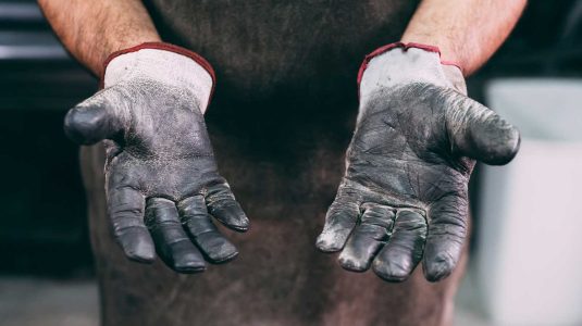 Close-up das mãos de um trabalhador com luvas de vaqueta desgastadas, evidenciando a necessidade diária da proteção das mãos no ambiente de trabalho industrial.