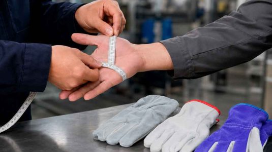 profissional medindo a circunferência da palma da mão de um trabalhador com fita métrica para definir o tamanho de luva adequado, com três luvas de segurança sobre uma mesa metálica
