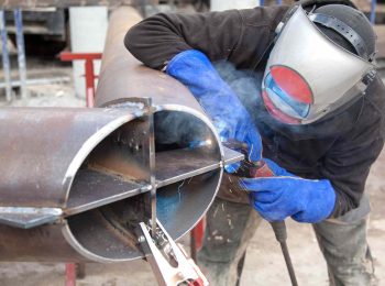 Soldador profissional equipado com máscara e luvas de proteção demonstra os constantes riscos na soldagem ao trabalhar em uma grande tubulação de aço industrial.