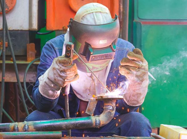 saude-dos-soldadores Um operário de solda está trabalhando em um ambiente industrial, usando equipamentos de proteção, como máscara facial, luvas grossas e roupa de trabalho. Ele segura um tubo metálico com uma chama elétrica, gerando faíscas durante o processo de soldagem.