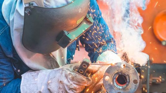 Close de um soldador profissional protegido com máscara de solda gerando uma nuvem de fumos de solda ao unir uma flange metálica.