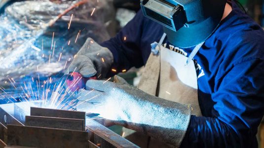 Soldador profissional usando, máscara de solda, luva de raspa e avental de raspa para se proteger das faíscas da solda em uma bancada de trabalho.