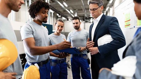 gerente em traje social conversa com operários em ambiente industrial para ilustrar como unir gerência e colaboradores na segurança do trabalho com foco em prevenção e uso de epis